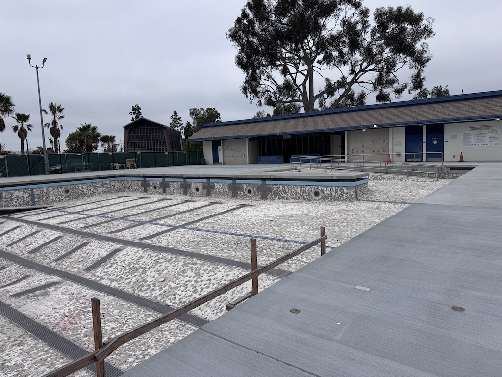 Clairemont Pool under renovation: empty basin awaiting plaster, new concrete deck completed, pool building in background.